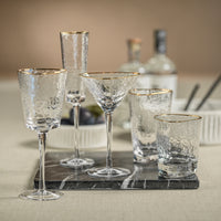 Set of clear glassware with gold rims including wine glasses, martini glasses, and tumblers on a marble tray.