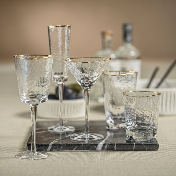Set of clear glassware with gold rims including wine glasses, martini glasses, and tumblers on a marble tray.
