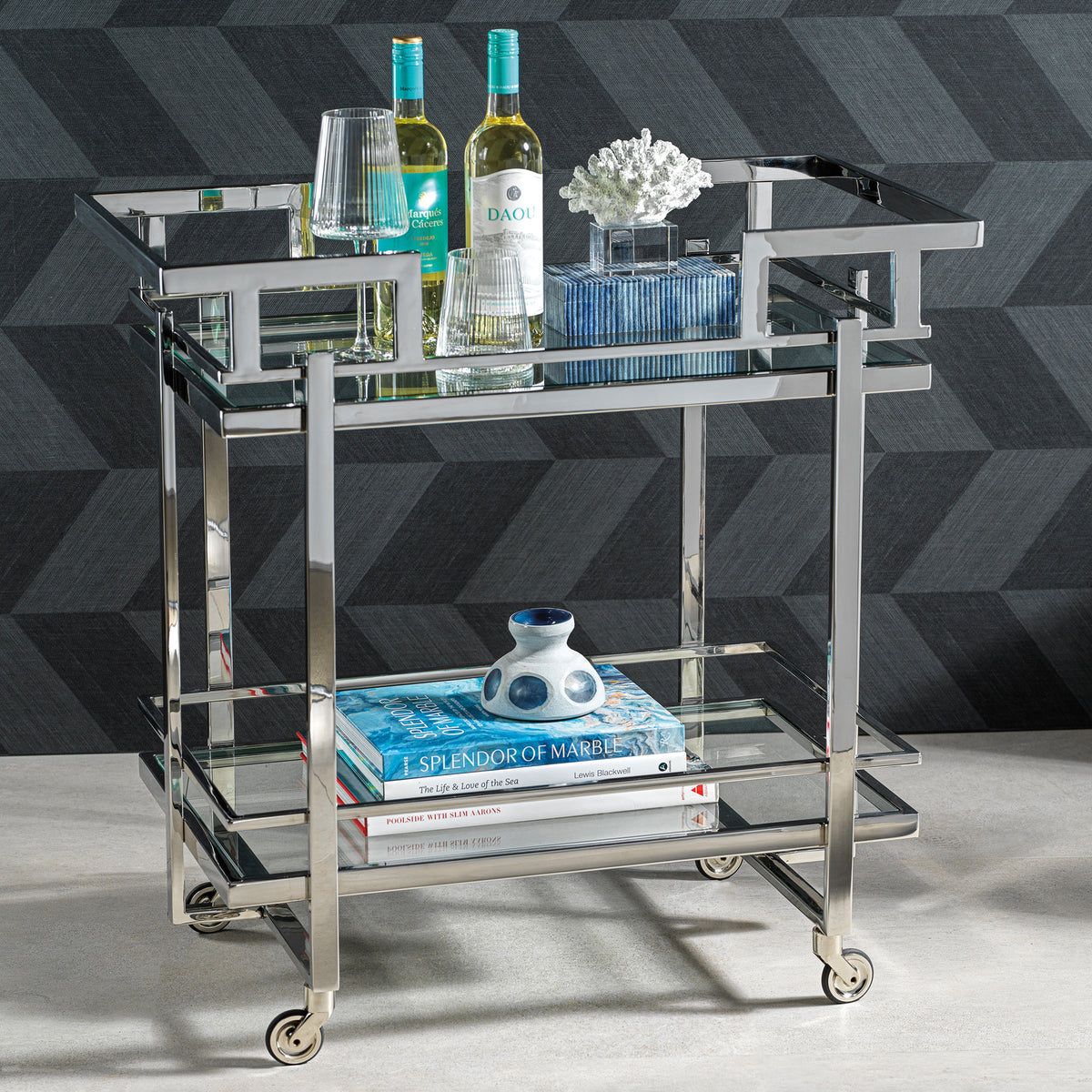 Glass bar cart with bottles, glasses, and books against a geometric patterned wall.