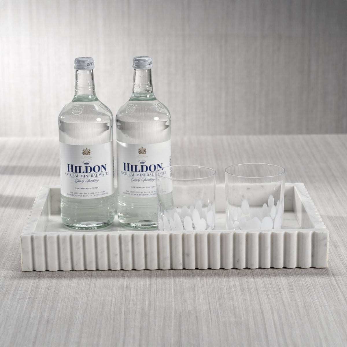 Two bottles of Hildon water on a marble tray with glasses against a neutral background
